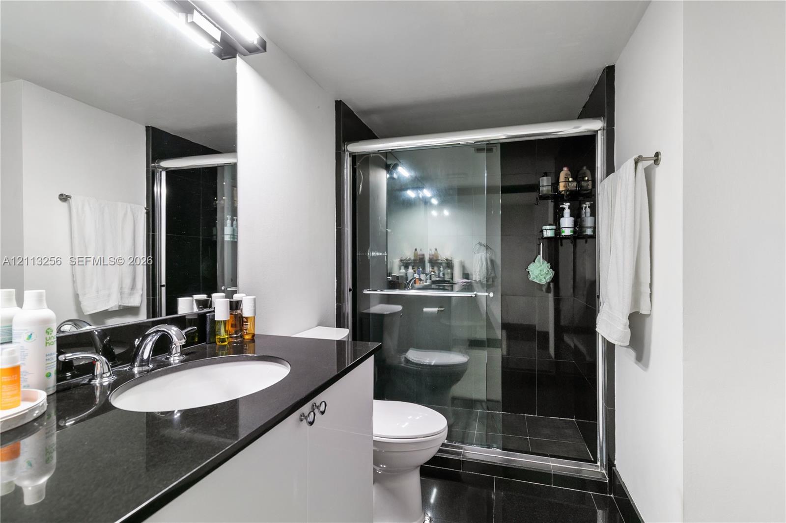 Bathroom featuring a dark stone countertop, dual sinks, white cabinetry, a glass-enclosed shower with dark tile surround, and black tiled flooring