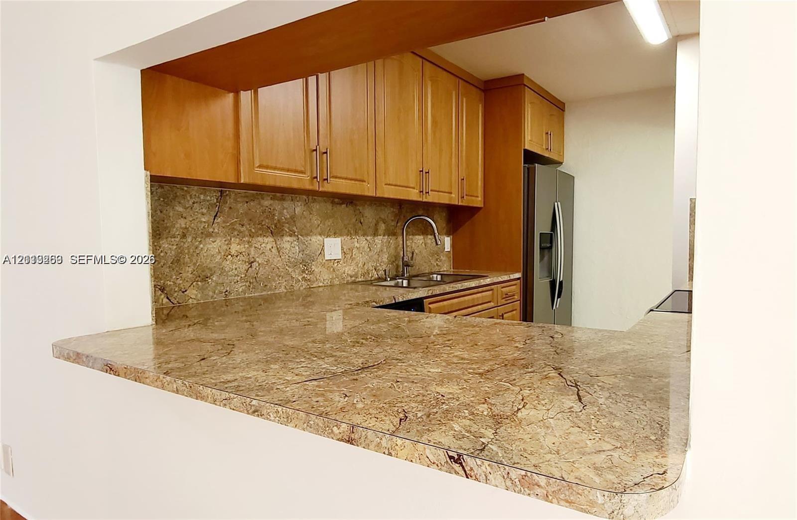 Kitchen featuring wood cabinetry, stone countertops, stone backsplash, stainless steel sink with gooseneck faucet, and stainless steel refrigerator