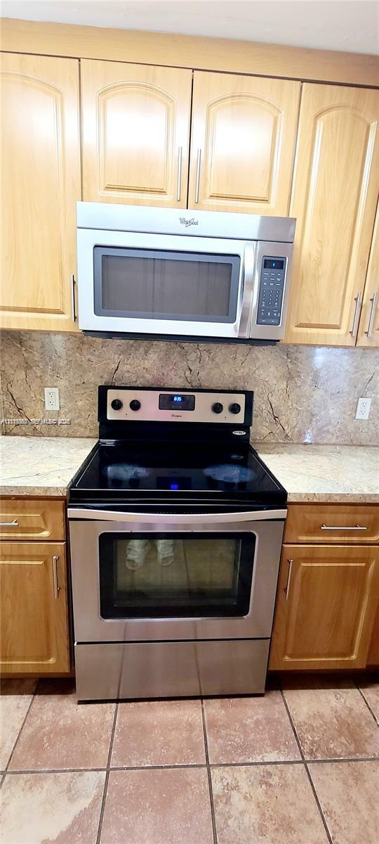 Light wood-finish cabinetry, stainless steel over-the-range microwave, electric range with black cooktop, stone-pattern backsplash, and tile flooring