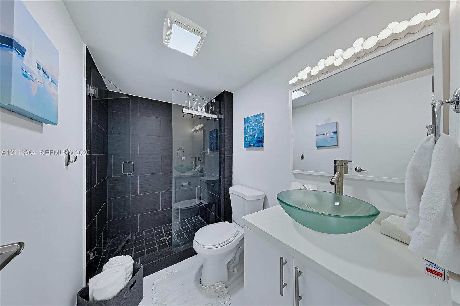 Bathroom featuring a walk-in shower with dark horizontal tile surround, glass vessel sink, white vanity, and wall-mounted light fixture