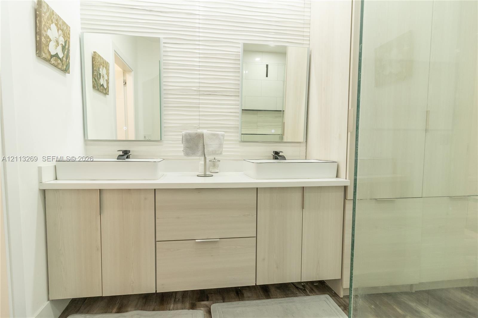 Double vanity featuring rectangular vessel sinks, light wood-tone cabinetry, a textured tiled wall accent, and frameless glass shower enclosure
