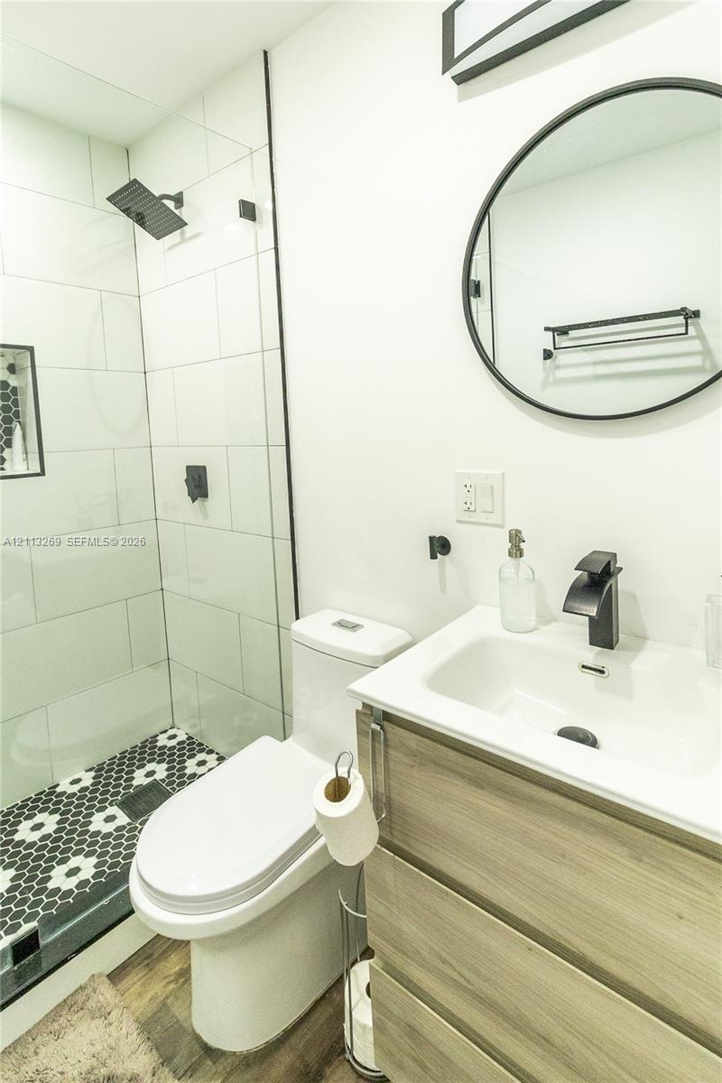Modern bathroom featuring a wood-finish vanity with a rectangular sink, matte black faucet, frameless glass-enclosed shower with oversized white wall tiles, and hexagonal mosaic floor tiling