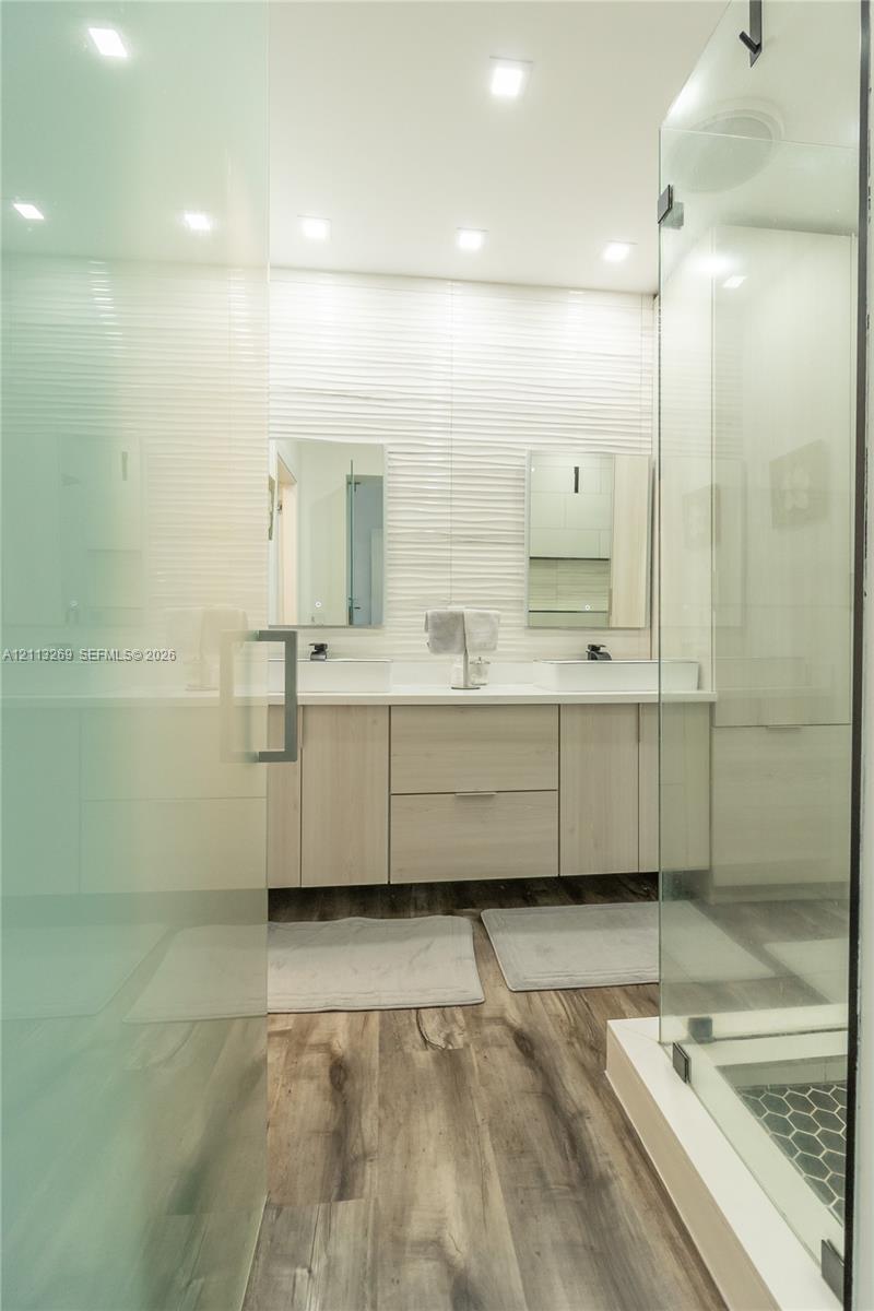 Dual-sink vanity with light wood-finish cabinetry, quartz countertops, textured tile backsplash, wood-finish flooring, and a glass-enclosed shower