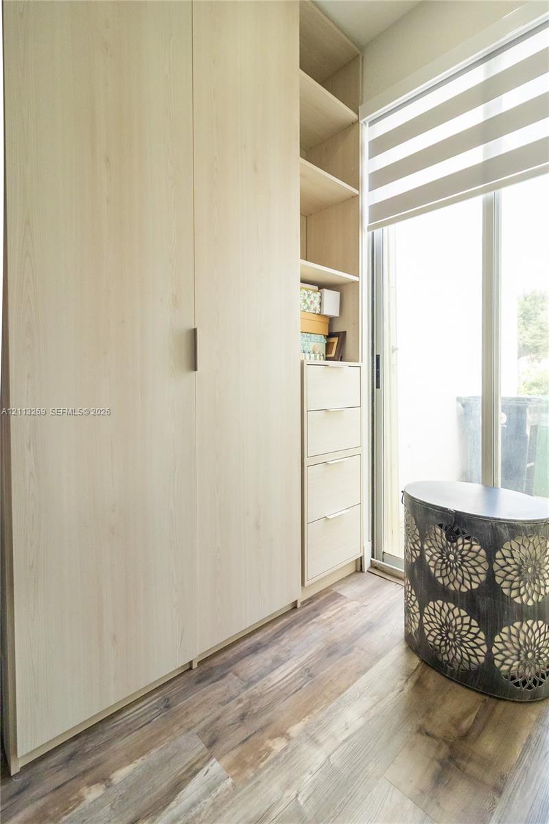 Light-toned wood cabinetry featuring integrated storage, open shelving, and drawer units with minimalist hardware, complemented by wood-finish flooring and a full-height glass door with installed roller shades