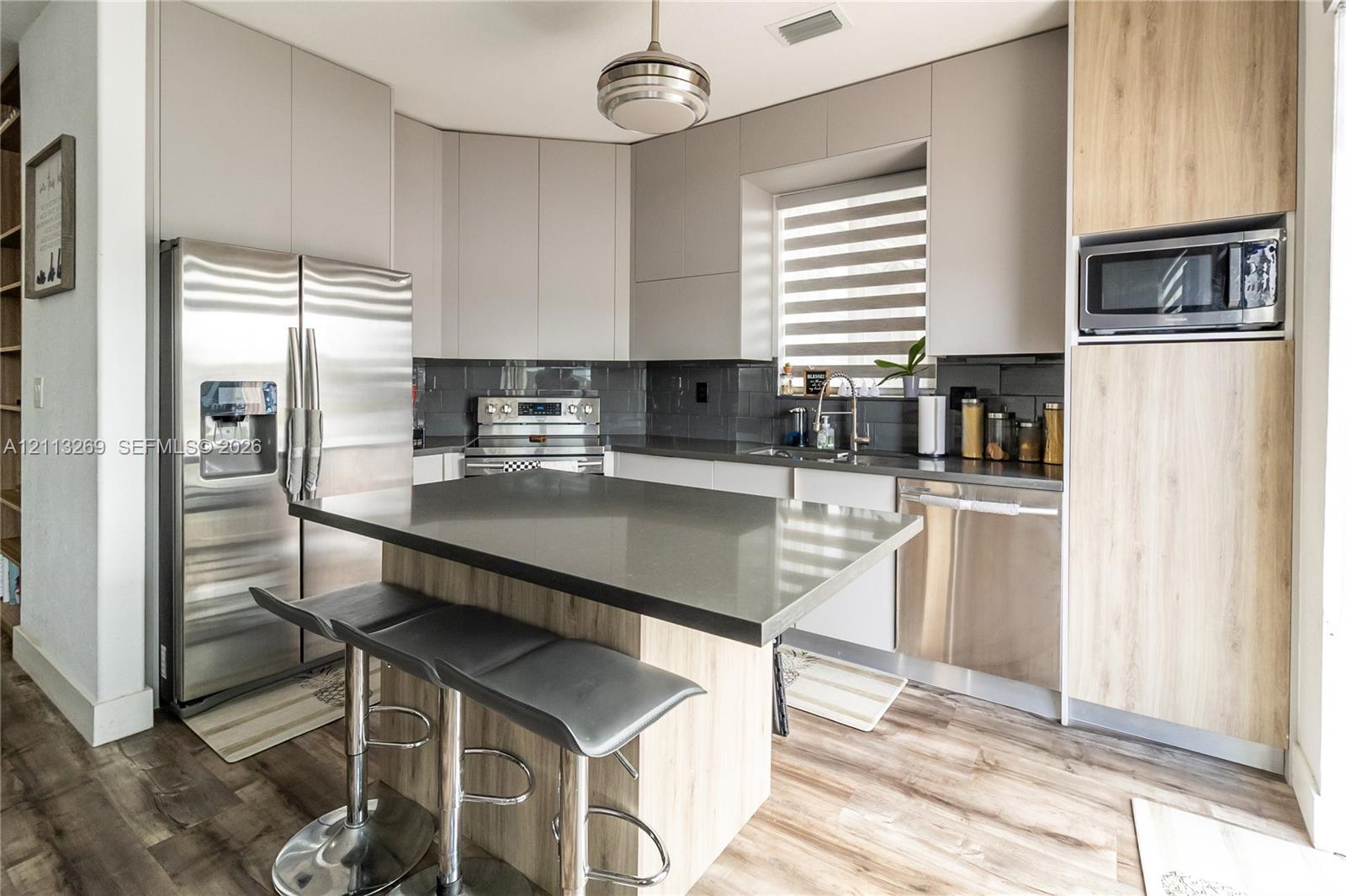 Modern kitchen featuring sleek flat-panel cabinetry in matte grey and wood-finish, stone-surfaced island, stainless steel appliances, and wood-finish flooring