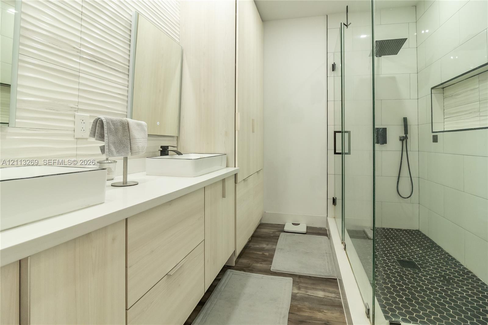 Dual-sink vanity with light wood-finish cabinetry, white solid-surface countertops, textured wall tile, frameless glass shower enclosure, and dark hexagon floor tile
