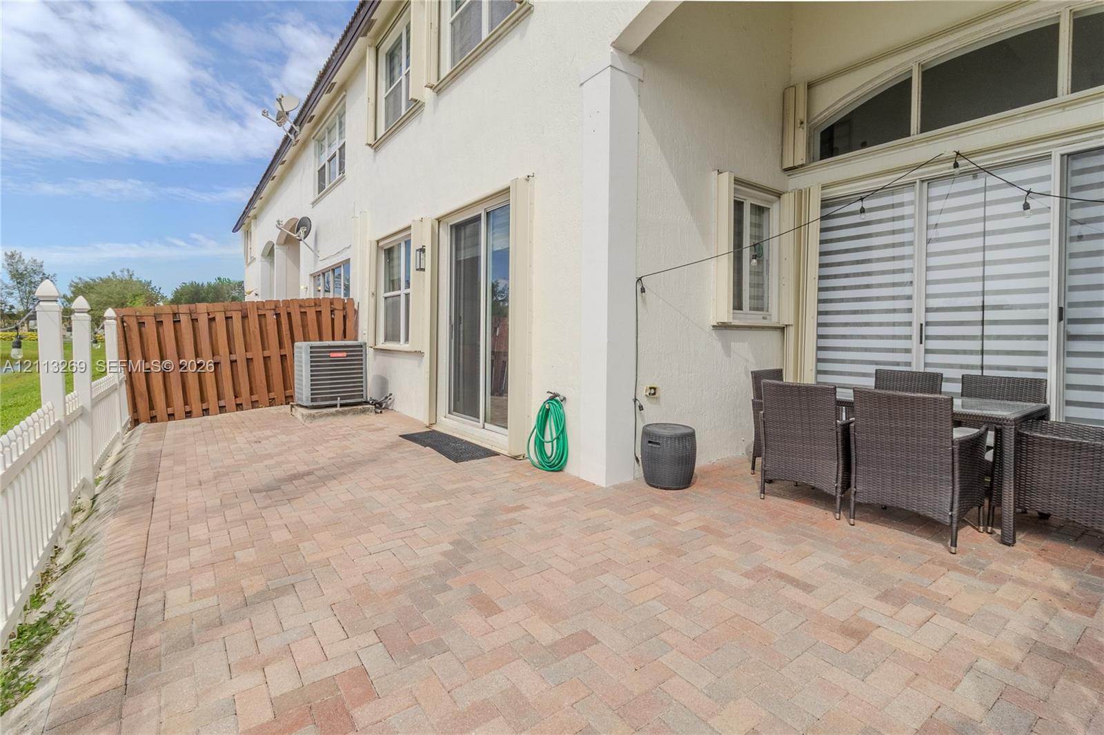 Rear patio featuring herringbone-patterned brick pavers, dual sliding glass door entry points, stucco exterior cladding, and white vinyl privacy fencing