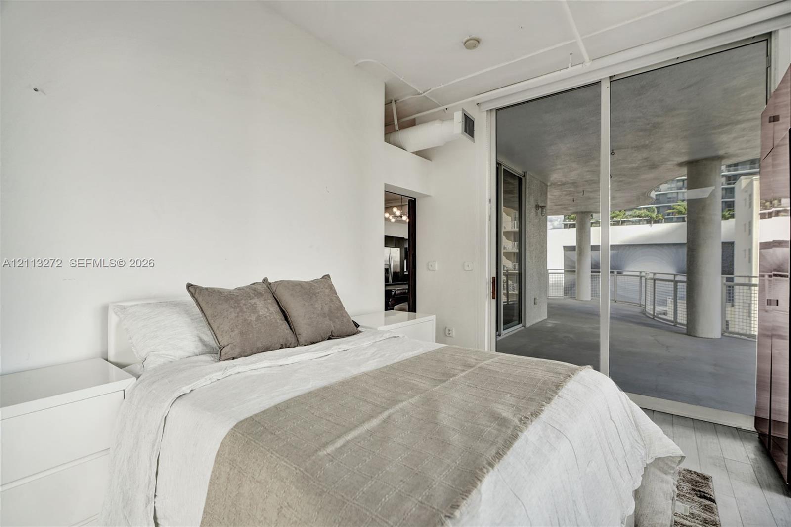 Floor-to-ceiling sliding glass door, private covered balcony with concrete pillars, exposed ceiling ductwork, and white wood-finish flooring