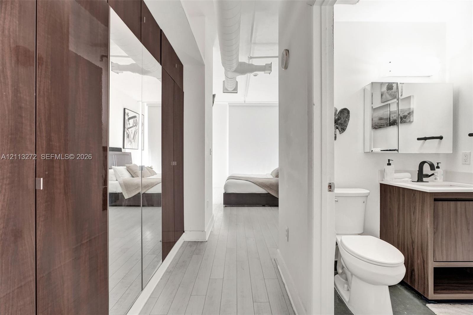 Wood-finish floor-to-ceiling cabinetry with mirrored panels, exposed overhead ductwork, wood-finish vanity cabinet with integrated sink, matte black faucet, and mirrored medicine cabinet
