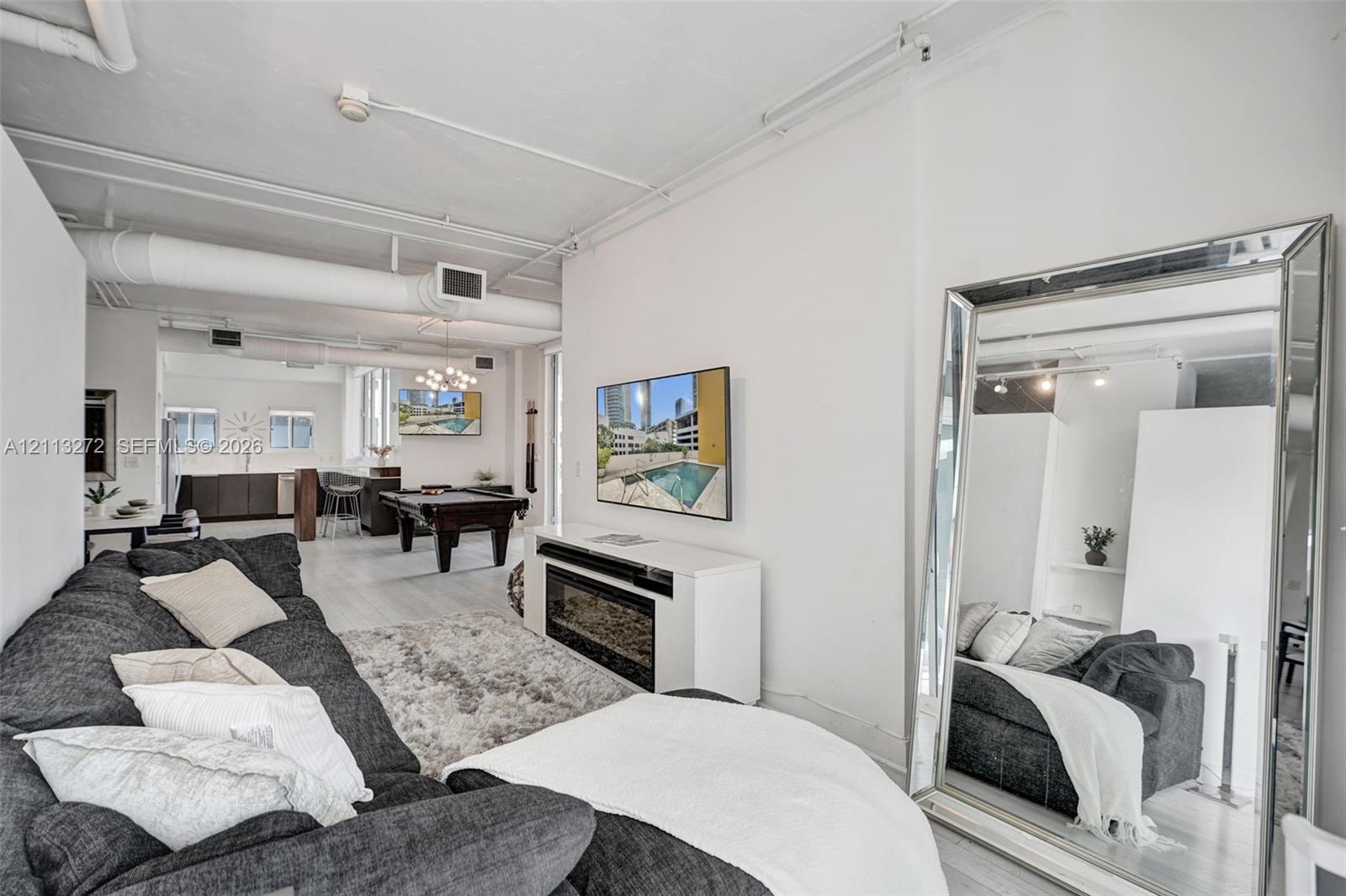 Open-concept interior featuring industrial-style exposed ductwork, white painted brick walls, wood-finish flooring, and a built-in wall-mounted electric fireplace