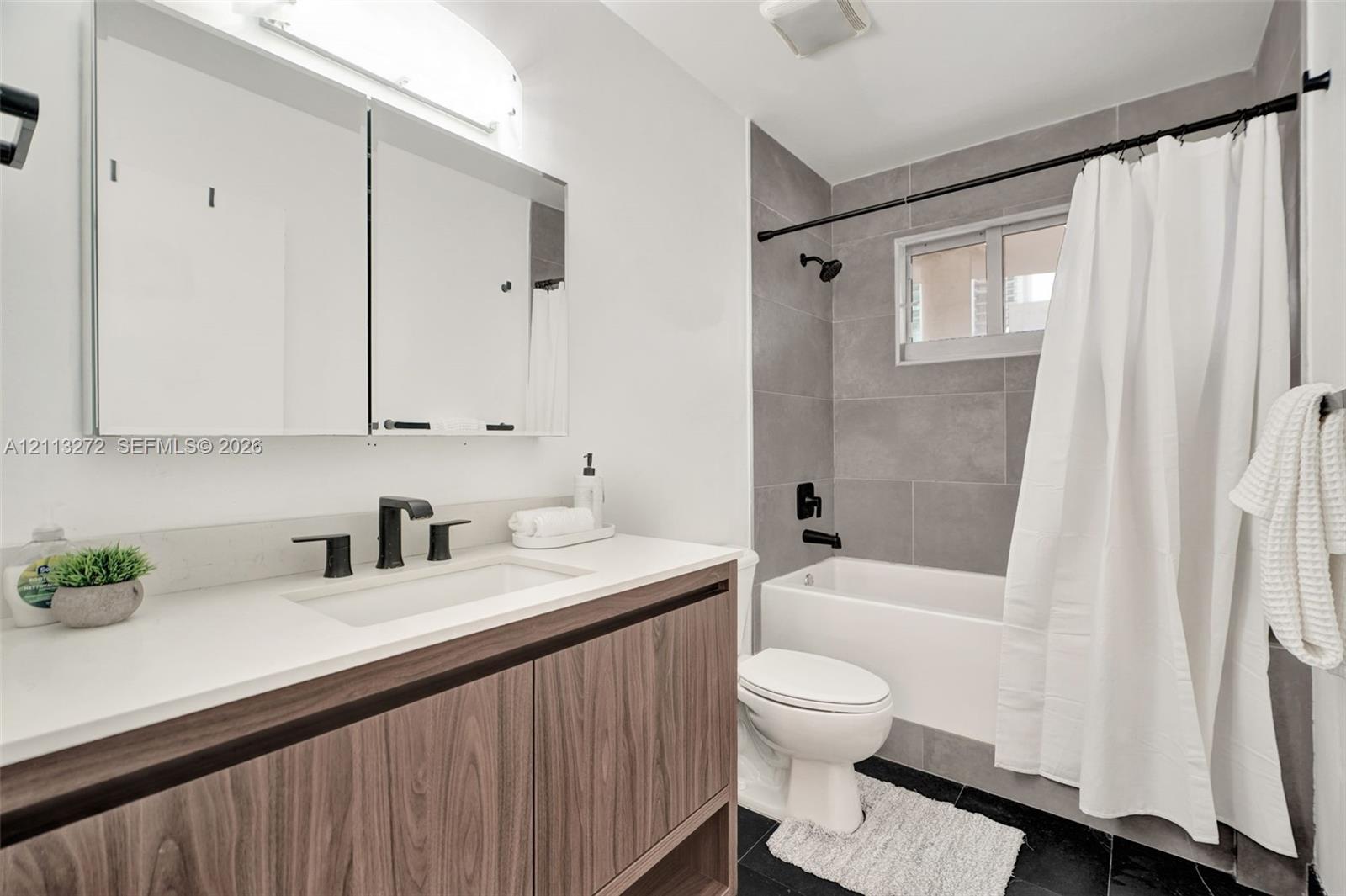 Bathroom featuring a wood-finish vanity with a white countertop, large mirrored medicine cabinet, matte black plumbing fixtures, and a tub-shower combination with grey tile surround