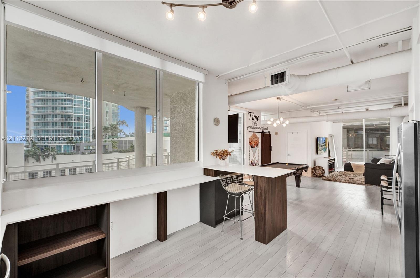 Expansive floor-to-ceiling windows, white-toned wood-finish flooring, industrial-style exposed ductwork, and built-in breakfast bar featuring a sleek white countertop and dark cabinetry