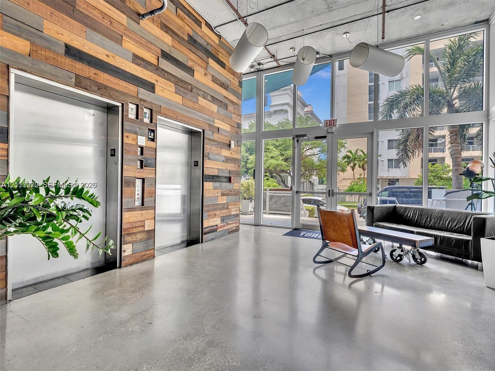 Industrial-style lobby featuring polished concrete flooring, a multi-tonal reclaimed wood accent wall, dual stainless steel elevator doors, floor-to-ceiling windows, and exposed ceiling infrastructure with cylindrical pendant lighting