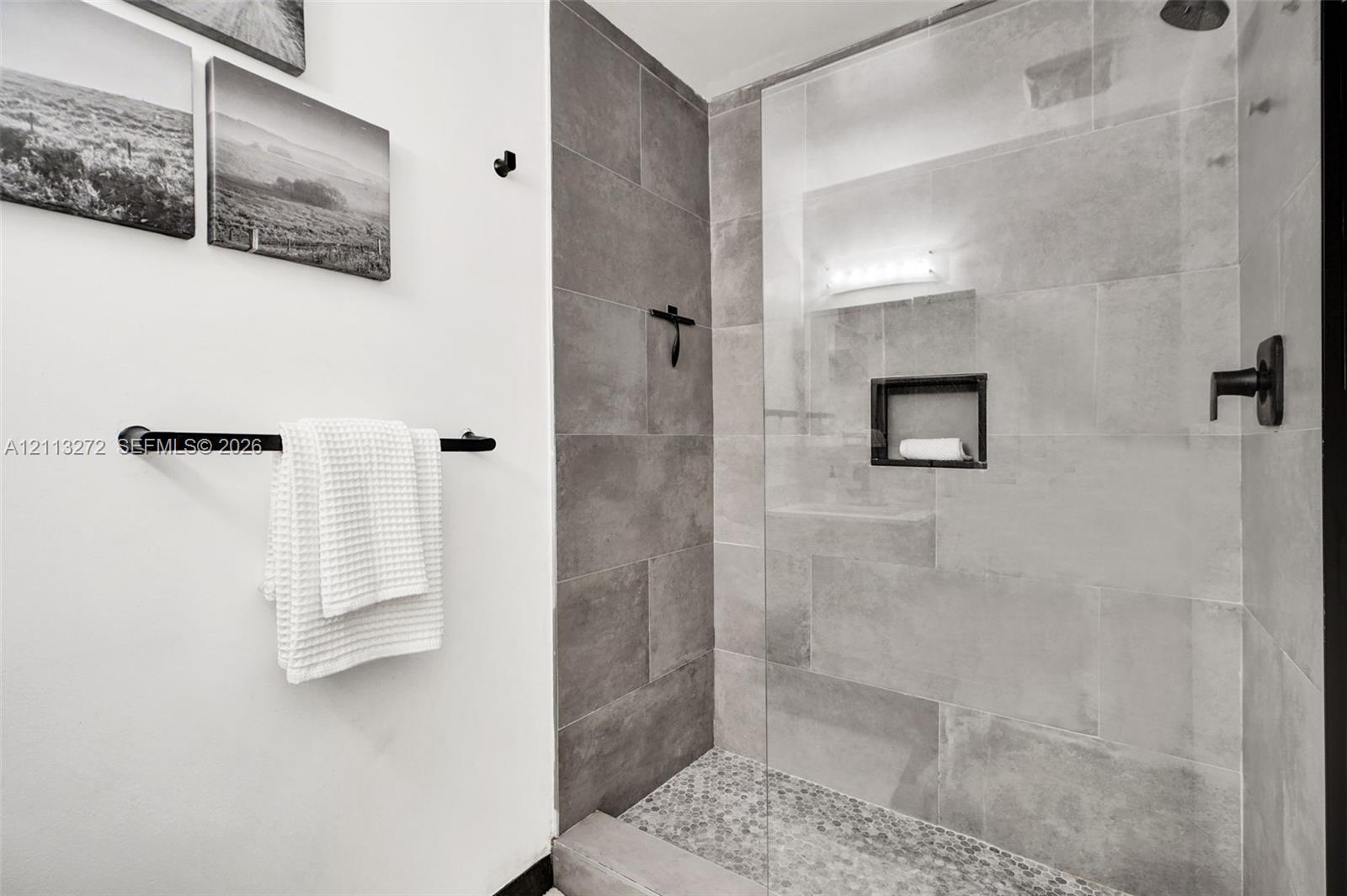 Walk-in shower featuring large-format grey tile surround, pebble-texture floor, frameless glass enclosure, built-in recessed wall niche, and matte black plumbing fixtures