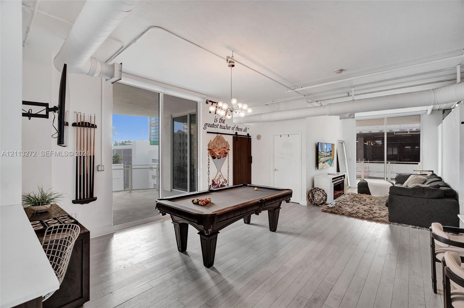 Open-concept industrial-style interior featuring high ceilings with exposed ductwork, wood-finish flooring, a multi-globe chandelier, and sliding glass doors providing access to the balcony