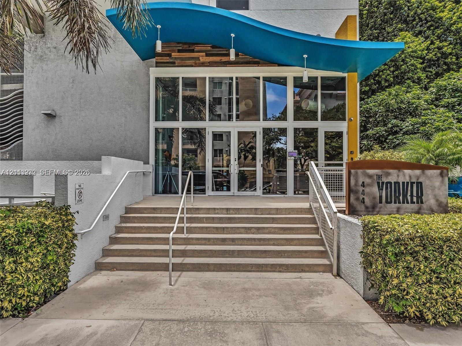Building entrance featuring a sweeping blue architectural canopy, floor-to-ceiling glass paneling, concrete steps, and white metal handrails