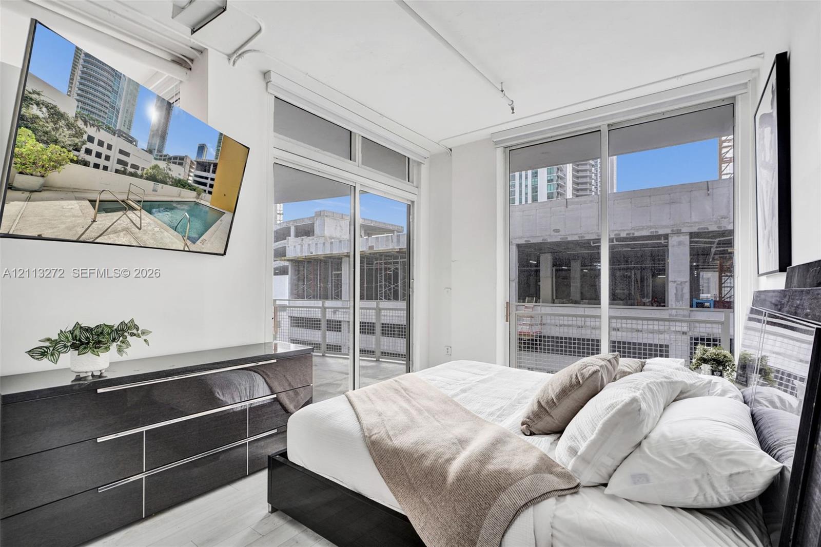 Floor-to-ceiling windows, sliding glass door with access to a balcony, high ceilings with exposed conduits, and light wood-finish flooring