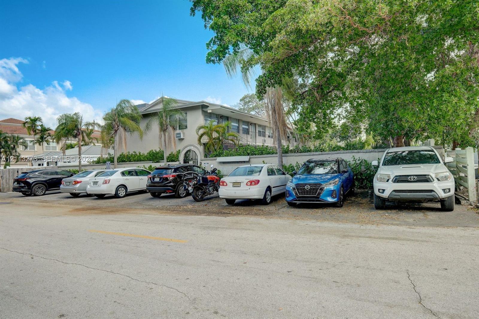 Multi-story exterior featuring smooth stucco siding, arched entryway, designated surface parking, and mature perimeter trees
