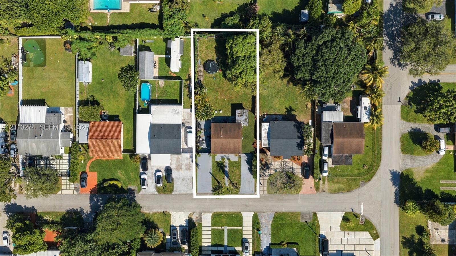 Aerial perspective of a residential lot featuring a concrete driveway, a central structure with a pitched roof, and an expansive rear yard with mature trees Aerial perspective of a residential lot featuring a concrete driveway, a central structure with a pitched roof, and an expansive rear yard with mature trees