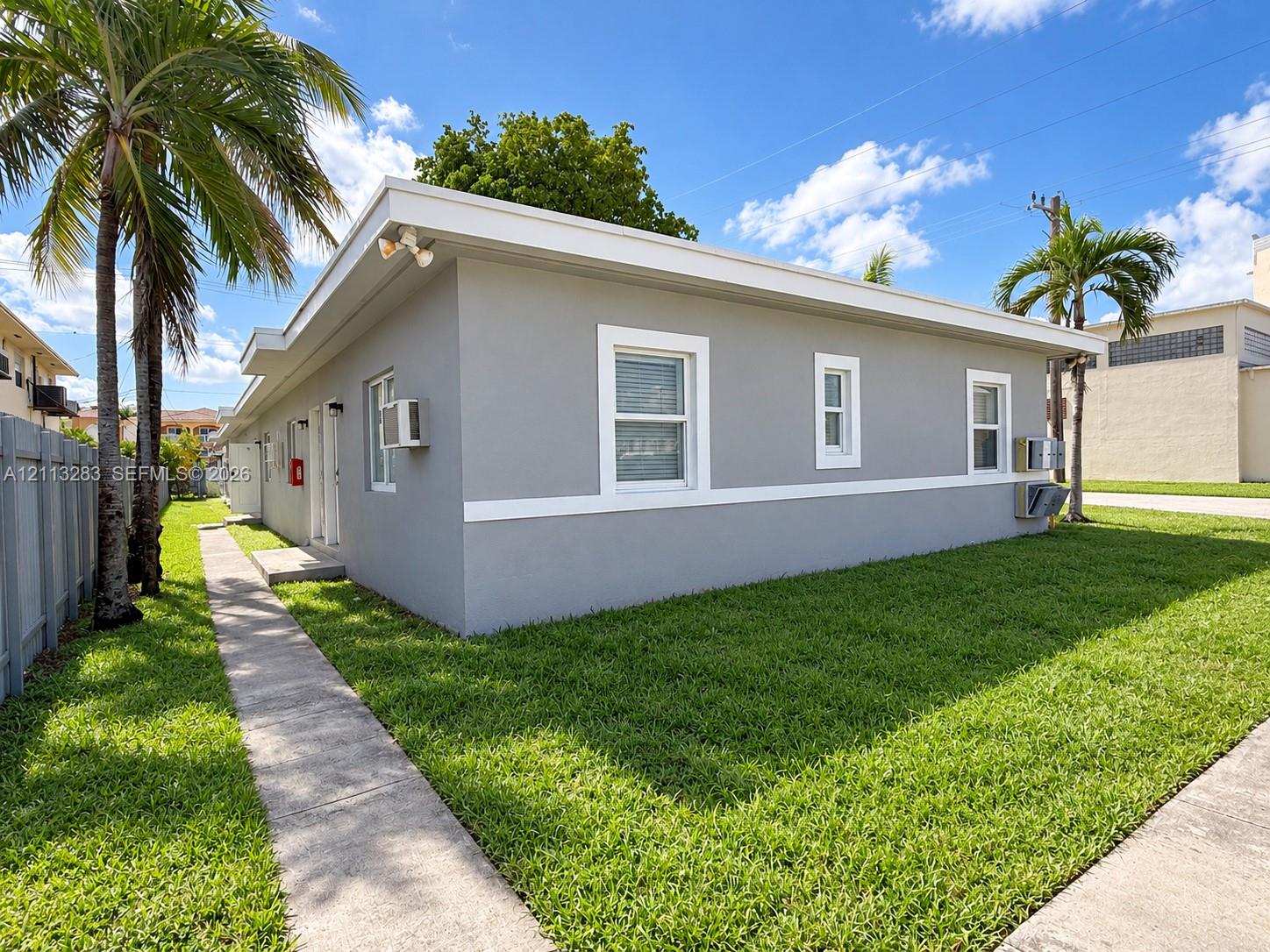 Single-story exterior featuring a neutral grey stucco facade, white trim detailing, flat roofline, and concrete walkway Single-story exterior featuring a neutral grey stucco facade, white trim detailing, flat roofline, and concrete walkway