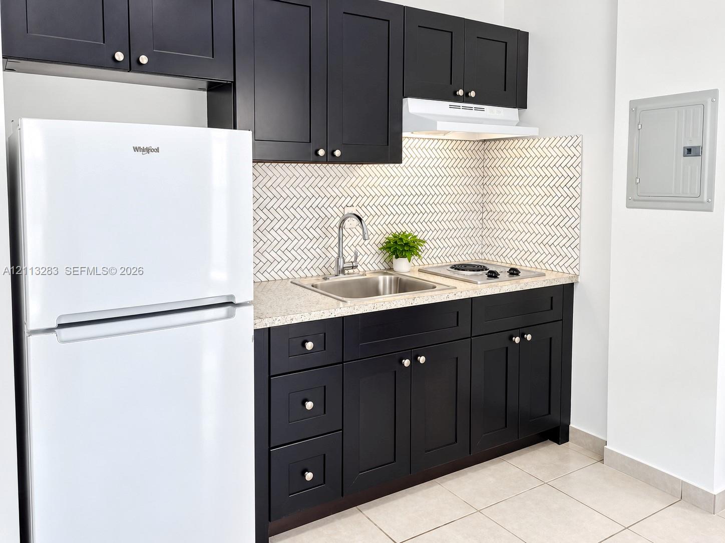 Kitchen featuring dark cabinetry, speckled stone-finish countertops, stainless steel sink with a high-arch faucet, herringbone-patterned tile backsplash, and a white refrigerator