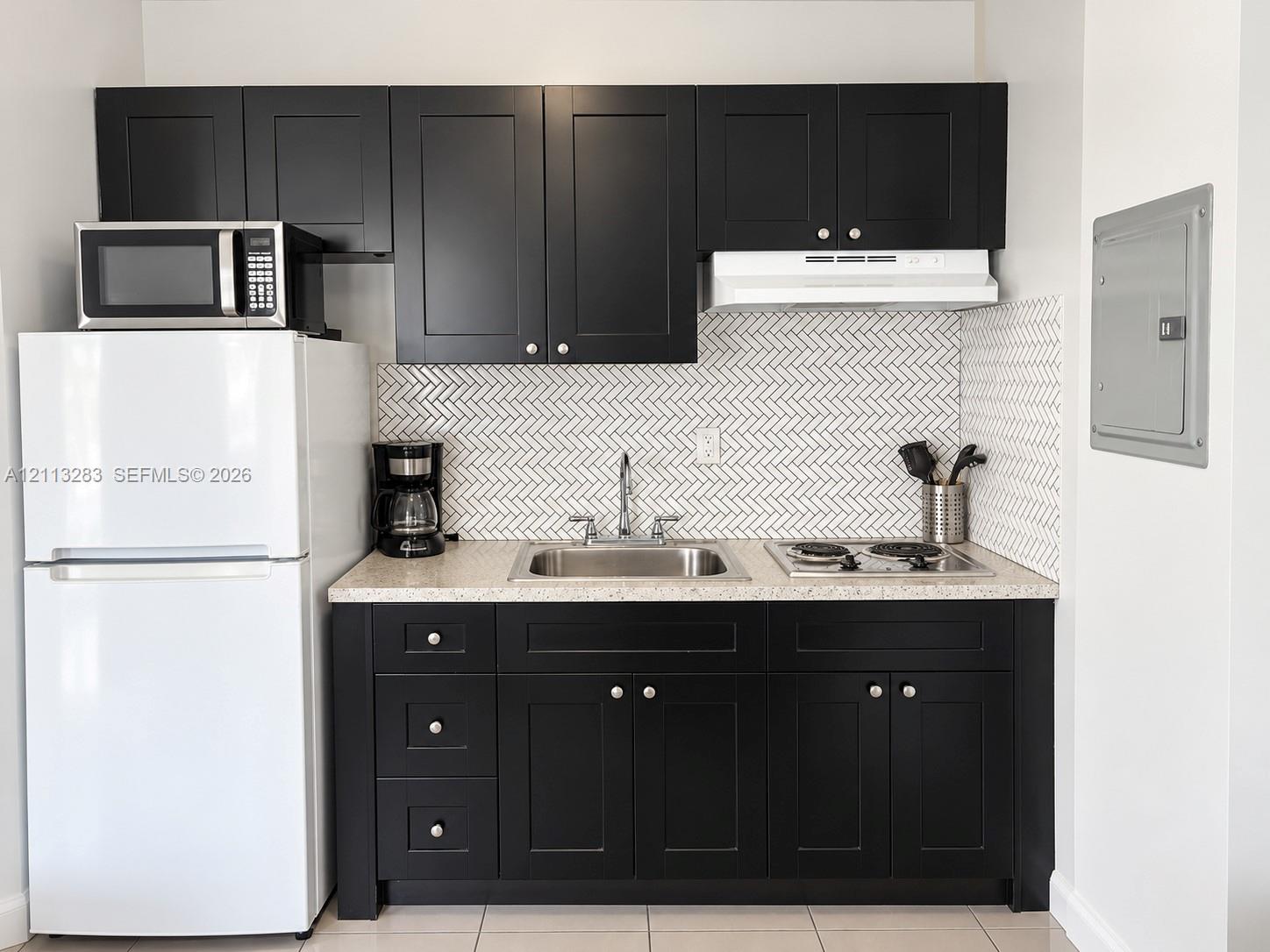 Kitchenette featuring matte black Shaker-style cabinetry, herringbone-patterned tile backsplash, speckled composite countertop, stainless steel sink with dual-handle faucet, and integrated two-burner electric cooktop