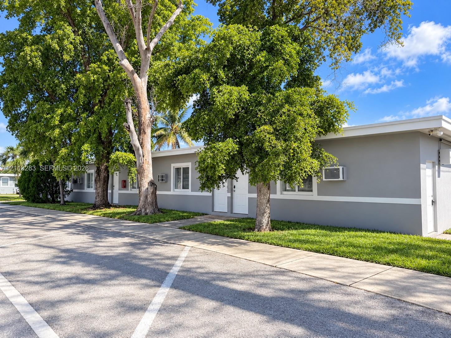 Single-story exterior featuring light gray painted masonry, white trim detailing, flat roofline, paved parking area, and mature trees Single-story exterior featuring light gray painted masonry, white trim detailing, flat roofline, paved parking area, and mature trees