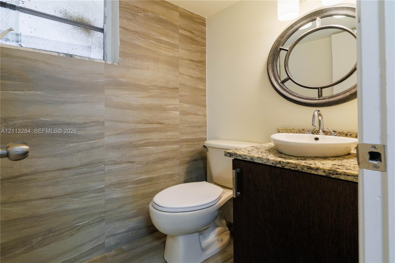 Vanity featuring a stone-look countertop, integrated oval vessel sink, and dark-finish cabinetry, complemented by wall-to-wall wood-tone tile and a circular decorative wall mirror