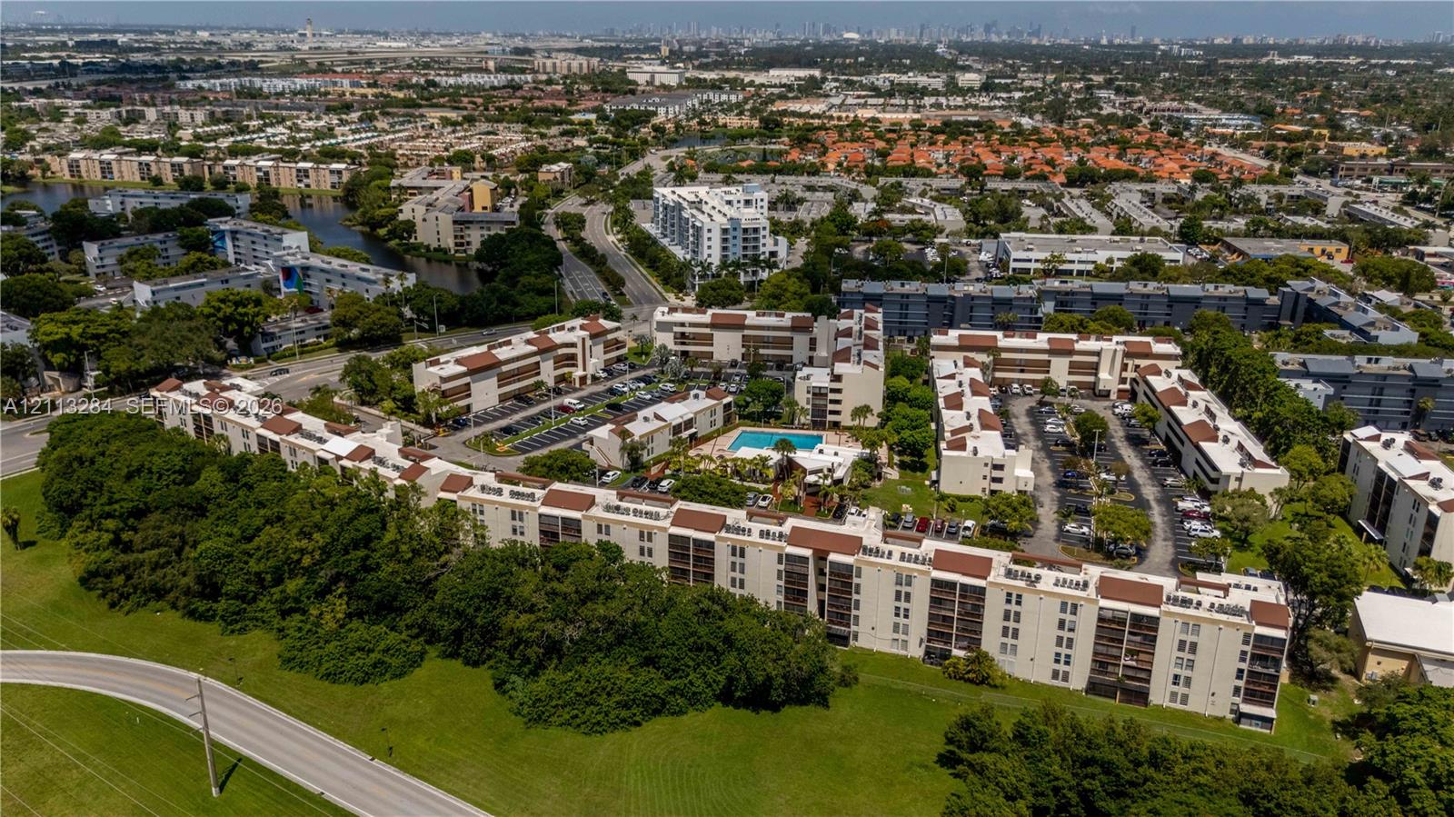 Aerial perspective of the condominium complex featuring multi-story structures with terra-cotta-toned roofing, an outdoor swimming pool, paved surface parking, and perimeter greenery