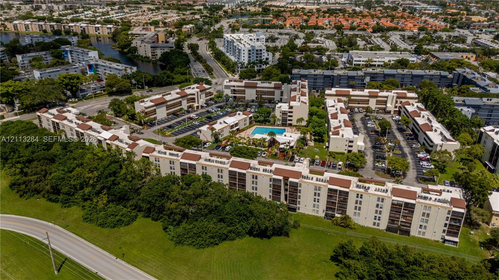 Aerial perspective of a multi-story residential complex featuring beige stucco facades, multi-pitched brown roofing, integrated parking areas, and a central outdoor swimming pool