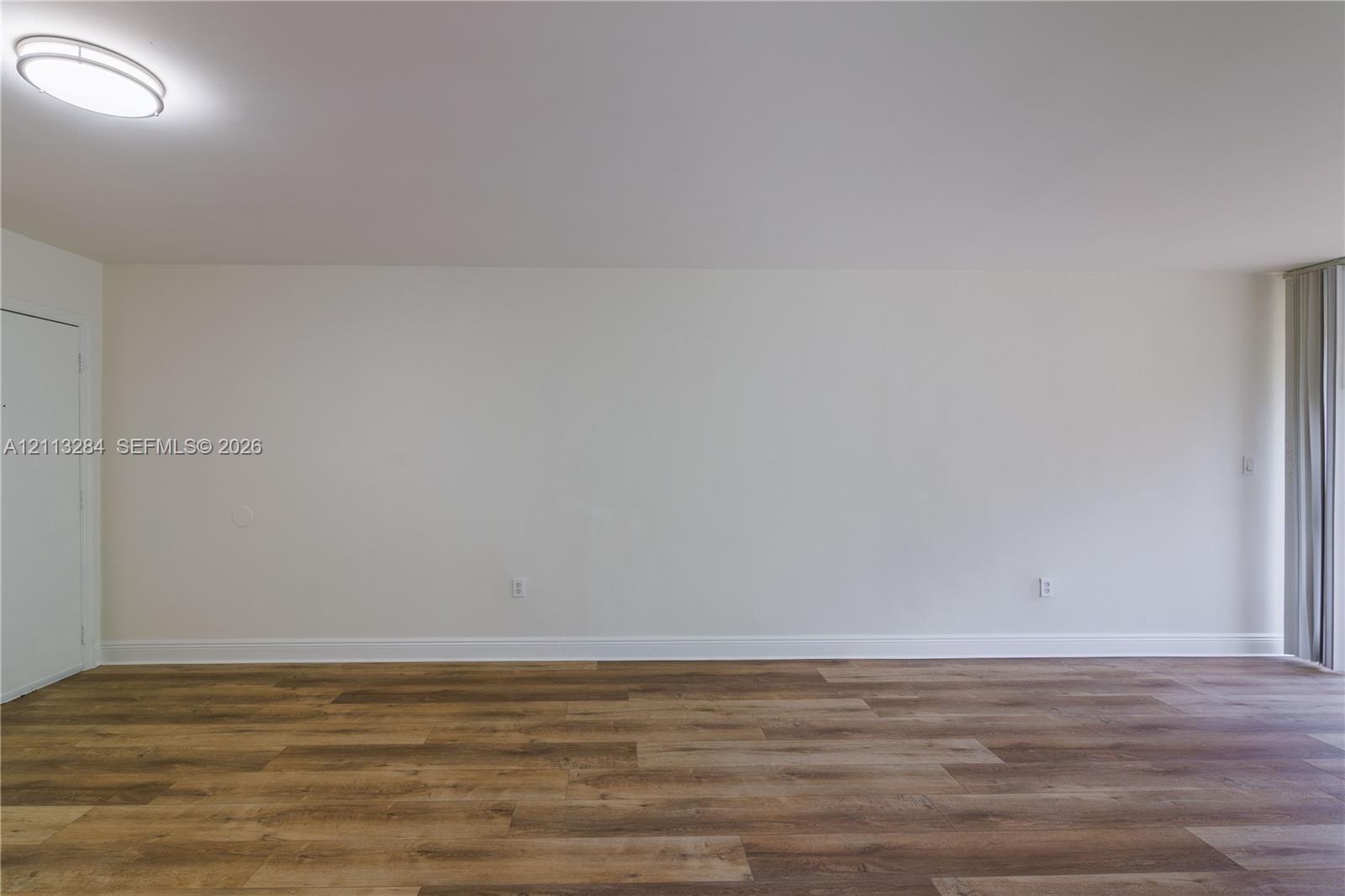 Interior space featuring wood-finish flooring, neutral wall tones, a ceiling-mounted flush light fixture, and vertical window blinds