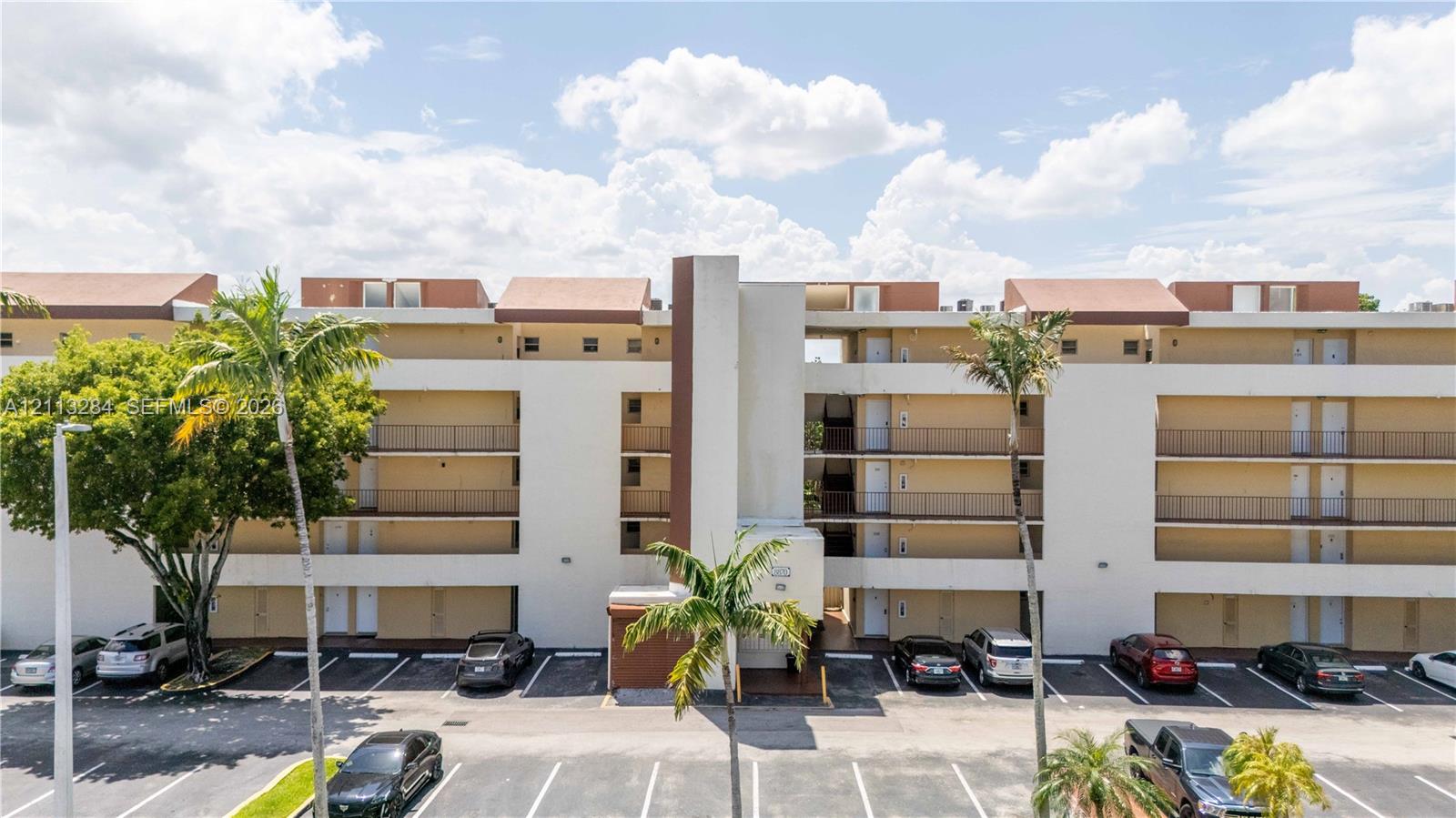 Multi-story residential building exterior featuring a neutral stucco facade, tiered exterior balconies, a prominent central architectural tower, and paved surface parking