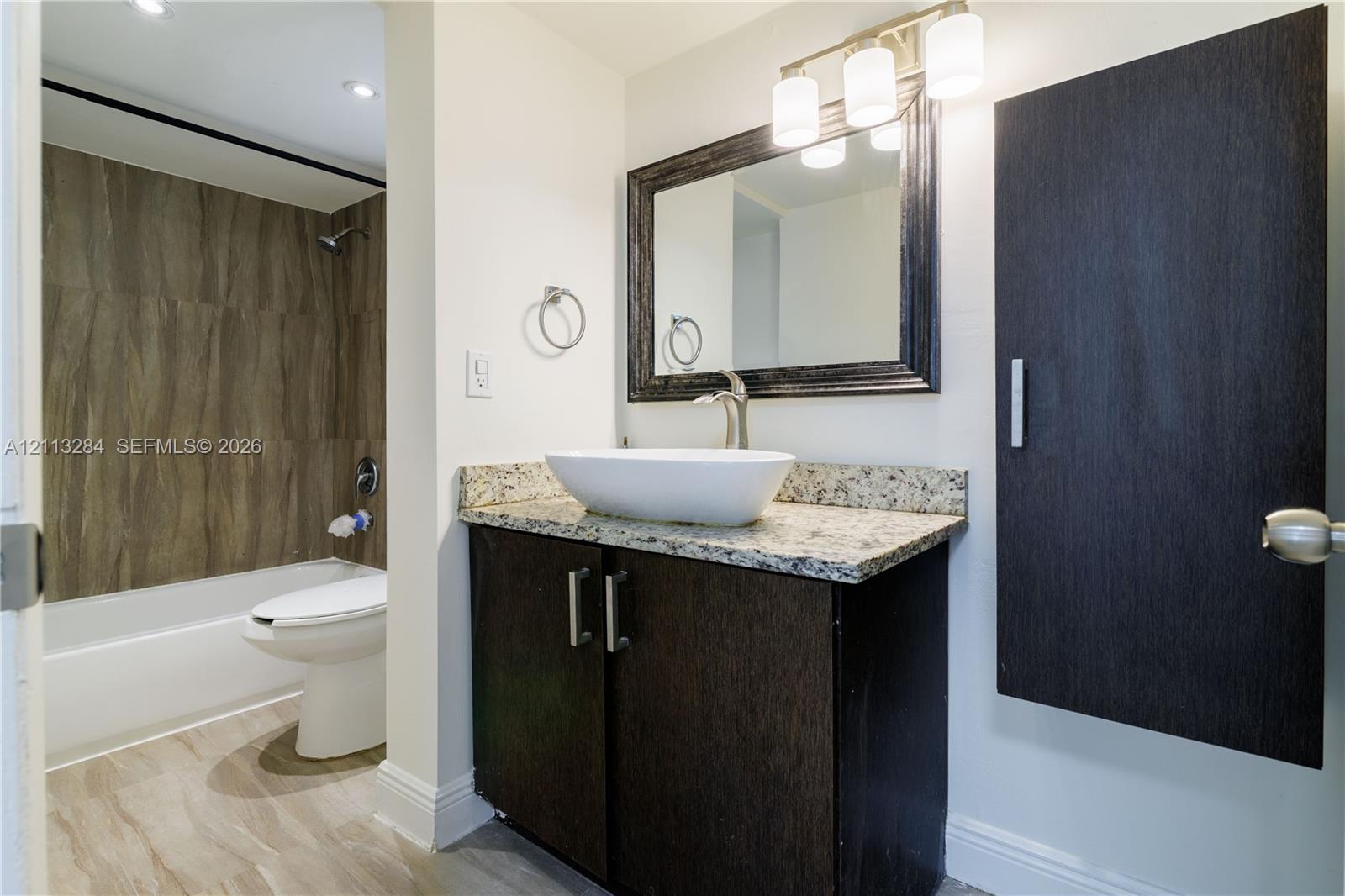 Bathroom featuring a dark wood-finish vanity with stone countertop, vessel sink, framed mirror with integrated light fixture, and a bathtub with tile surround