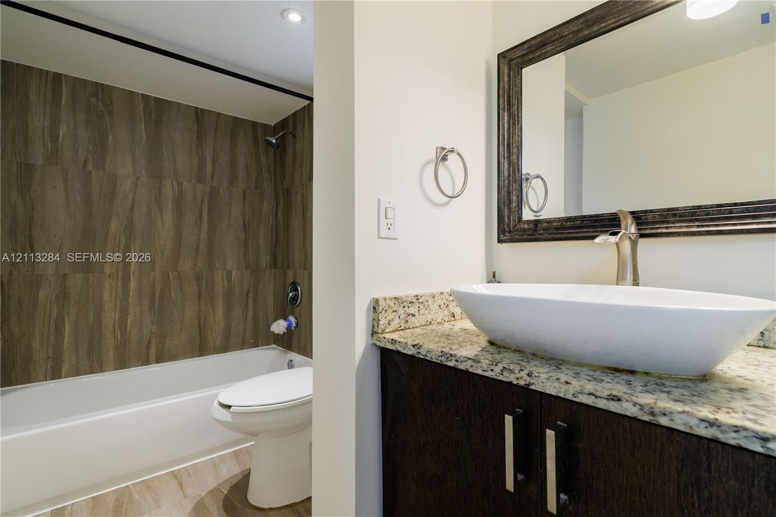 Bathroom featuring a dark wood-finish vanity with a stone countertop, an elongated vessel sink, metal faucet hardware, and a tub enclosure with full-height tile surround