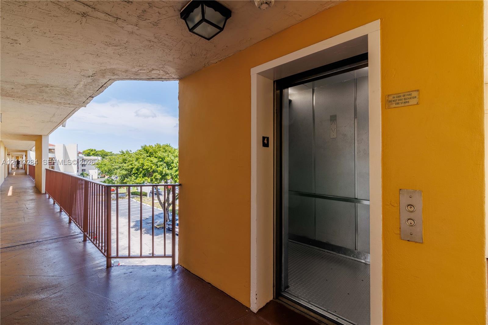 Elevator access featuring a metal-finish elevator cabin, painted concrete flooring, an overhead recessed light fixture, and an open-air corridor with metal railing