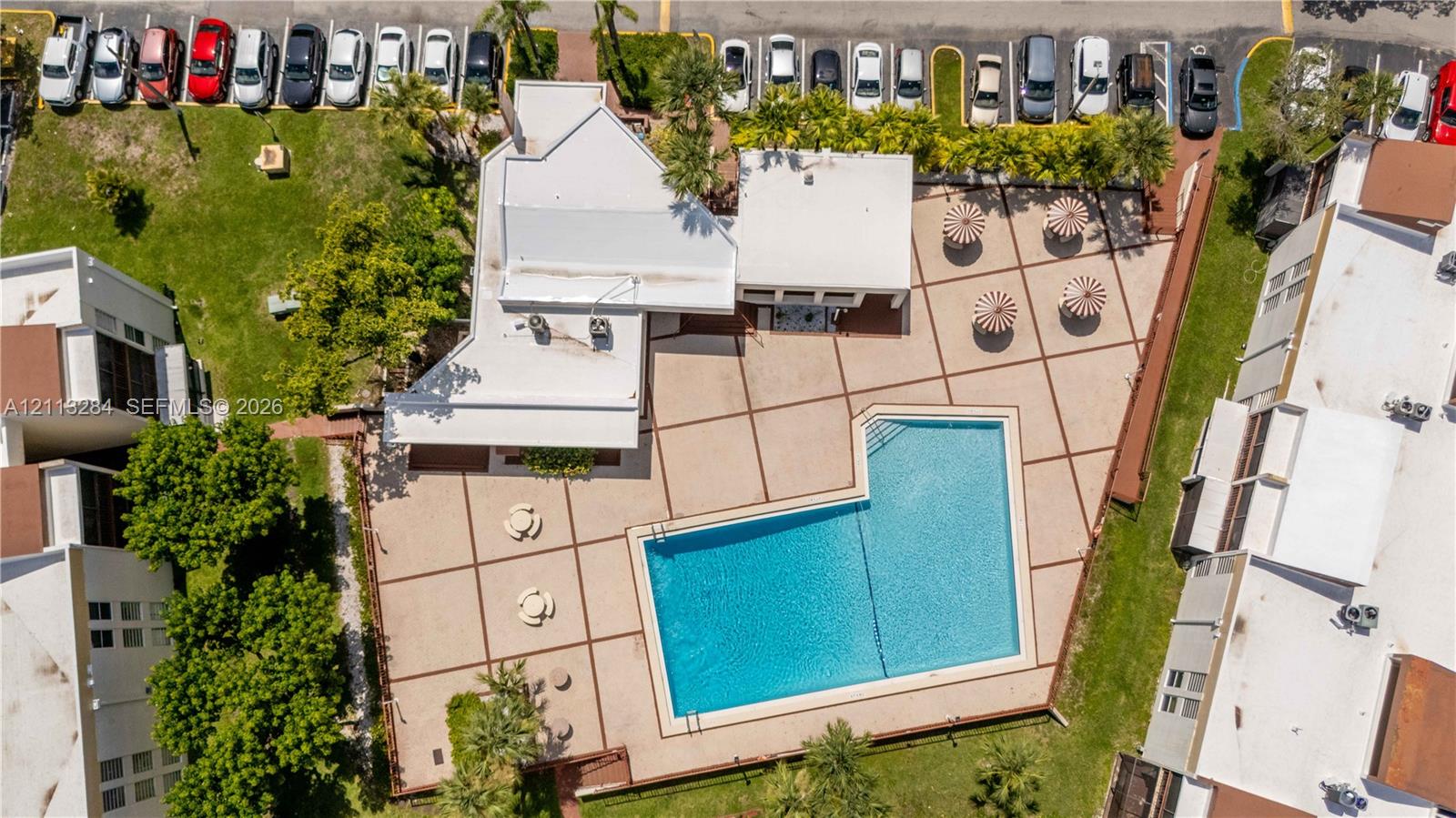 Aerial perspective of property featuring an expansive concrete pool deck, in-ground swimming pool, mature perimeter landscaping, and dedicated parking area