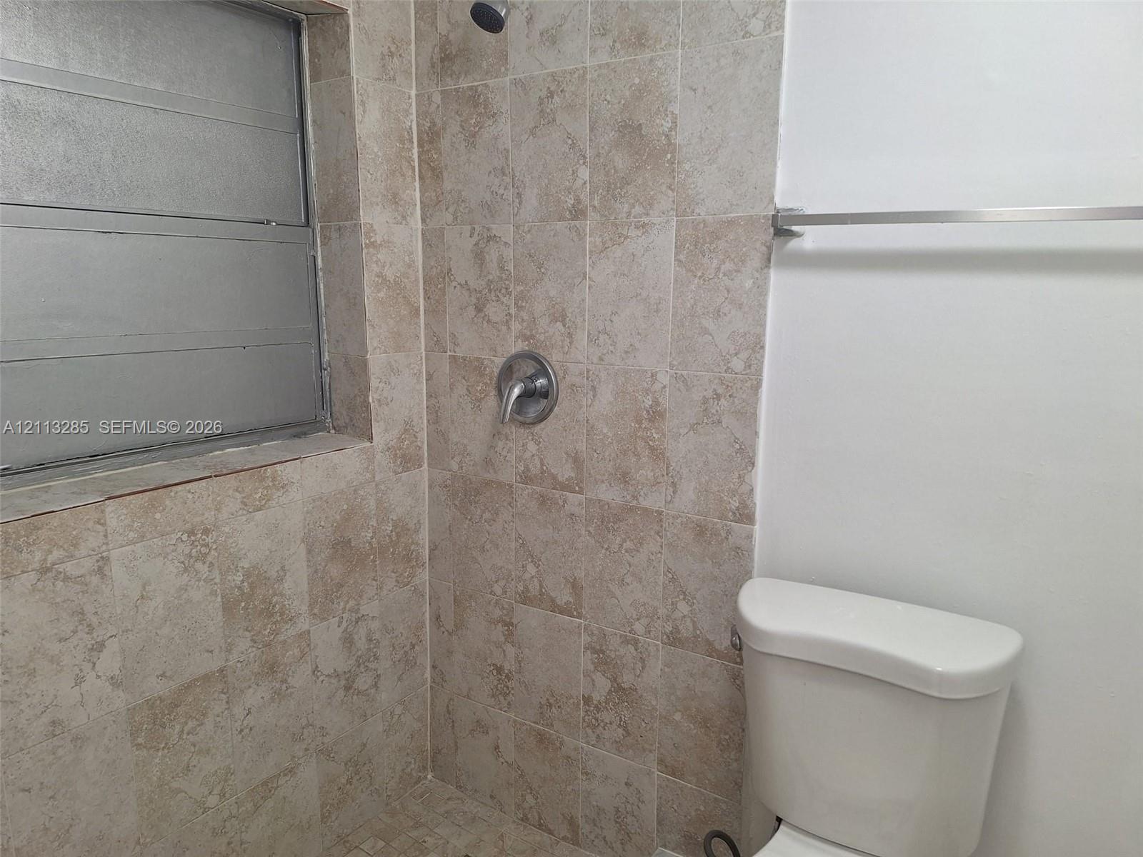Shower enclosure featuring textured tile walls, a stainless steel single-handle shower faucet, a louvered window, and a wall-mounted metal towel bar Shower enclosure featuring textured tile walls, a stainless steel single-handle shower faucet, a louvered window, and a wall-mounted metal towel bar