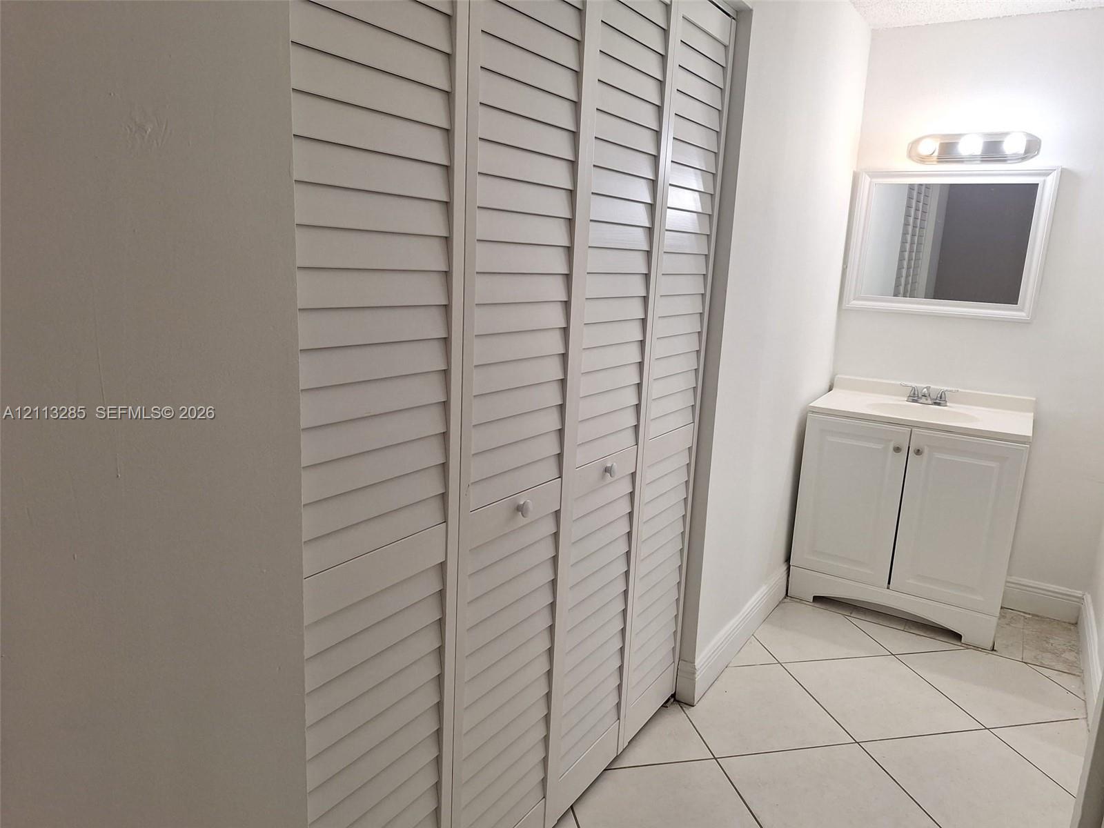 White louvered closet doors, vanity with integrated basin, polished light-colored tiled flooring, and wall-mounted vanity mirror with integrated lighting fixture White louvered closet doors, vanity with integrated basin, polished light-colored tiled flooring, and wall-mounted vanity mirror with integrated lighting fixture