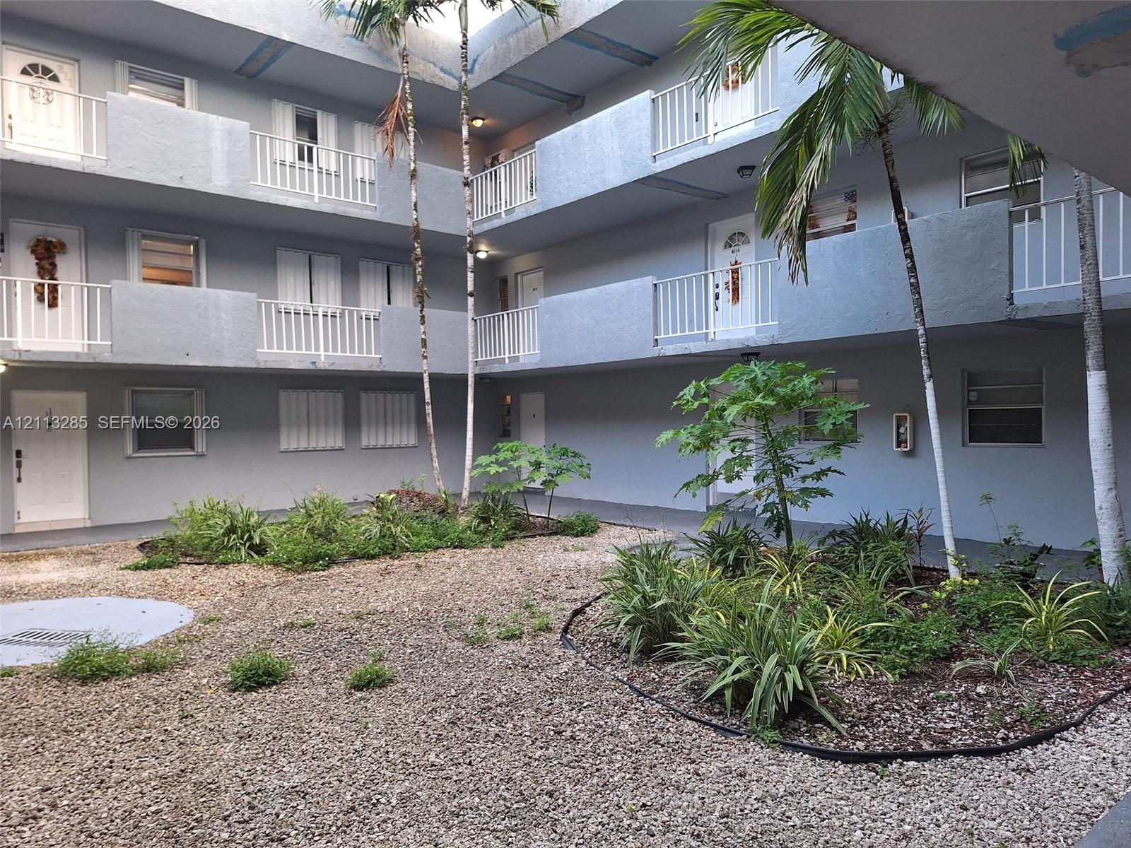 Multi-level exterior featuring white metal balcony railings, a central gravel courtyard, and mature palm trees