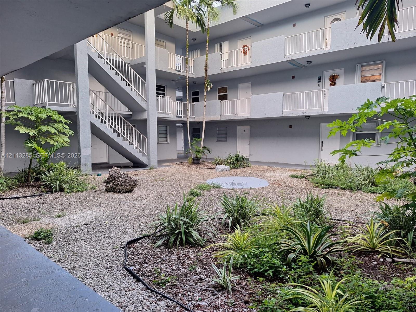 Multi-level structure featuring a gravel courtyard, palm trees, white metal railings, and exterior staircases Multi-level structure featuring a gravel courtyard, palm trees, white metal railings, and exterior staircases