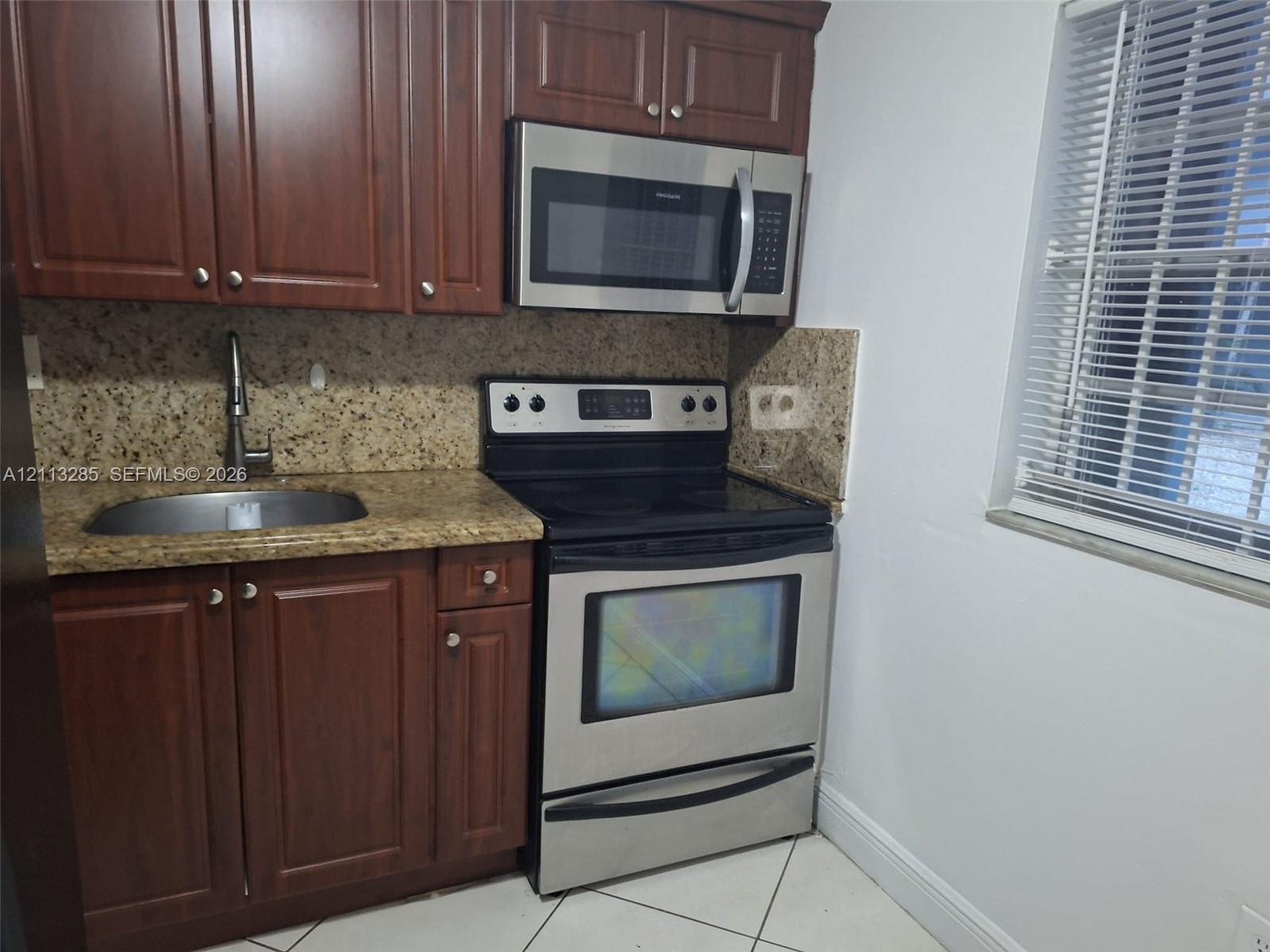 Kitchen area featuring dark-tone wood cabinetry, speckled stone countertops, stainless steel appliances, an undermount sink with a gooseneck faucet, and tile flooring Kitchen area featuring dark-tone wood cabinetry, speckled stone countertops, stainless steel appliances, an undermount sink with a gooseneck faucet, and tile flooring