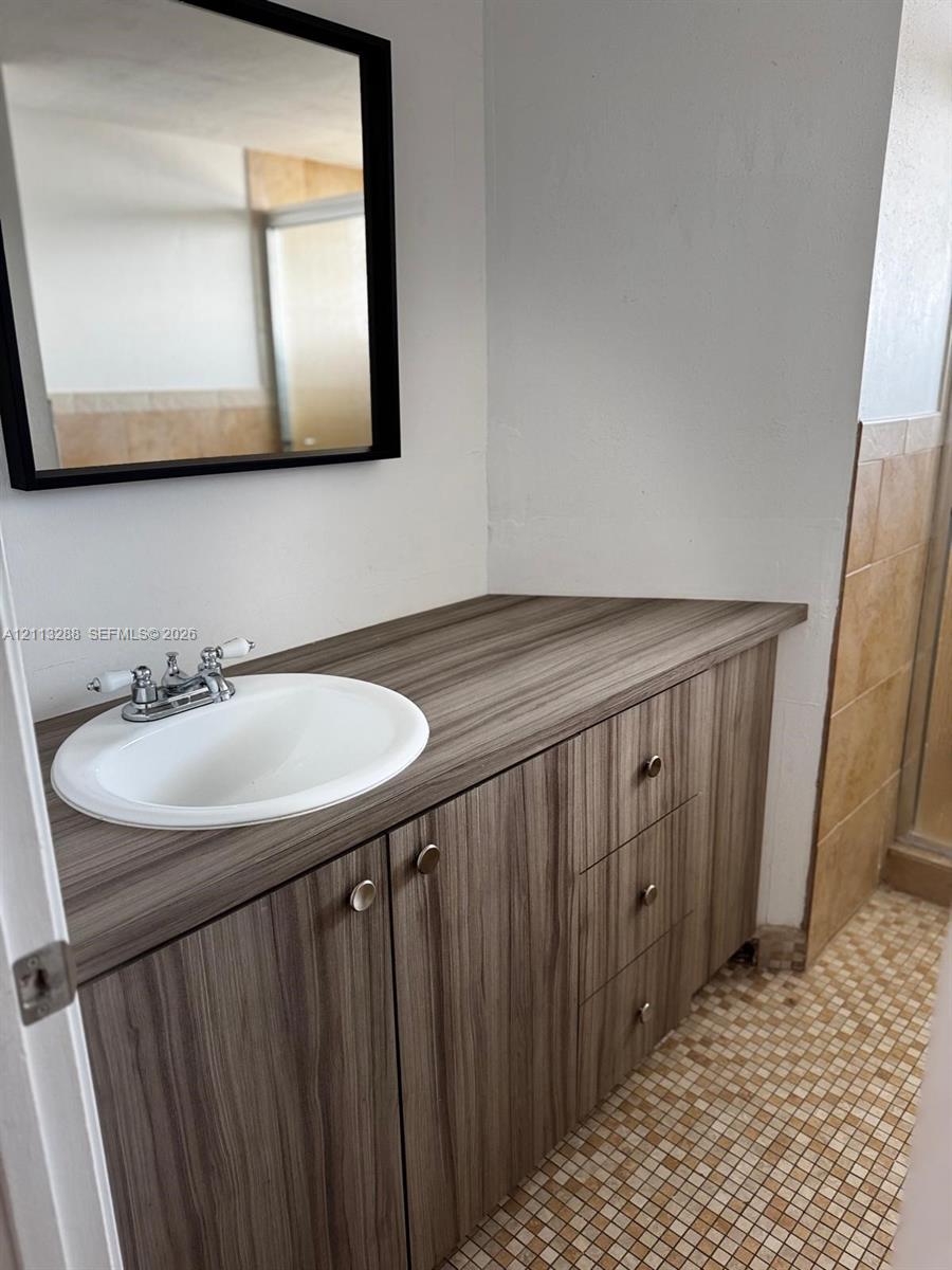 Vanity featuring wood-finish cabinetry, a drop-in sink, dual-handle chrome faucet, wall-mounted mirror with a black frame, and tiled flooring Vanity featuring wood-finish cabinetry, a drop-in sink, dual-handle chrome faucet, wall-mounted mirror with a black frame, and tiled flooring