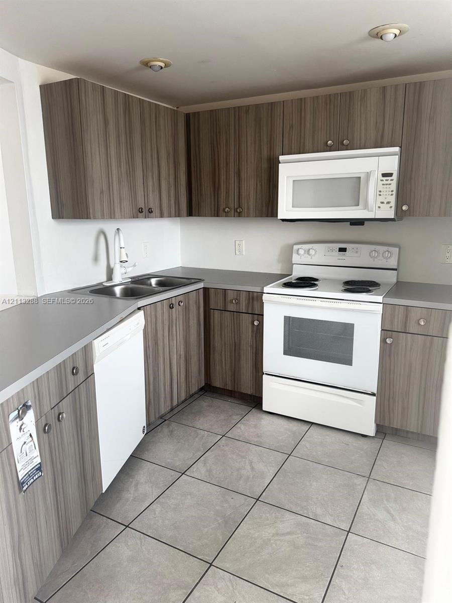 Kitchen featuring wood-finish cabinetry, neutral solid-surface countertops, large-format tile flooring, a dual-basin stainless steel sink, and white appliances Kitchen featuring wood-finish cabinetry, neutral solid-surface countertops, large-format tile flooring, a dual-basin stainless steel sink, and white appliances