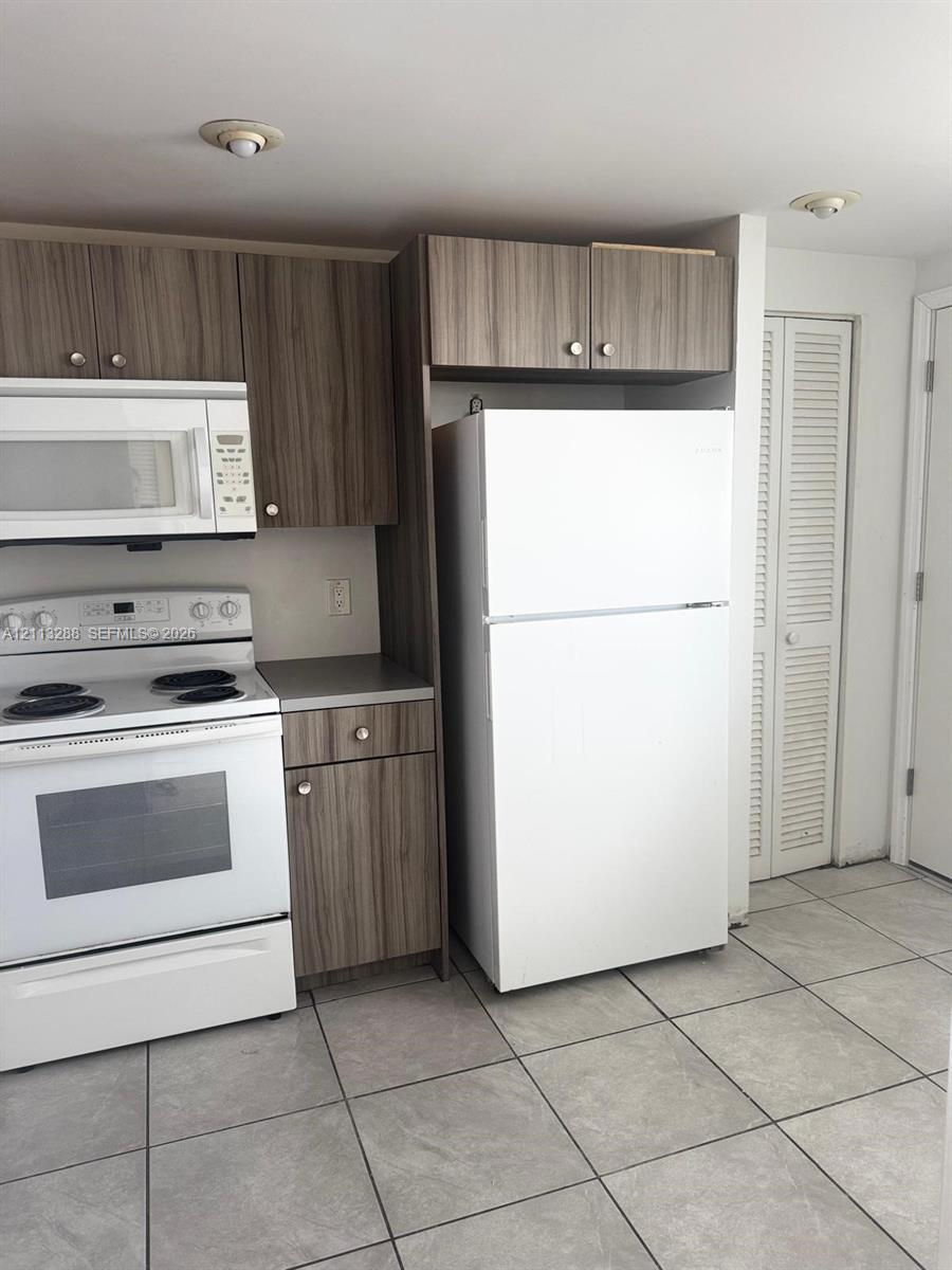 Kitchen featuring wood-finish cabinetry, white appliances, and large-format tile flooring with louvered closet doors Kitchen featuring wood-finish cabinetry, white appliances, and large-format tile flooring with louvered closet doors
