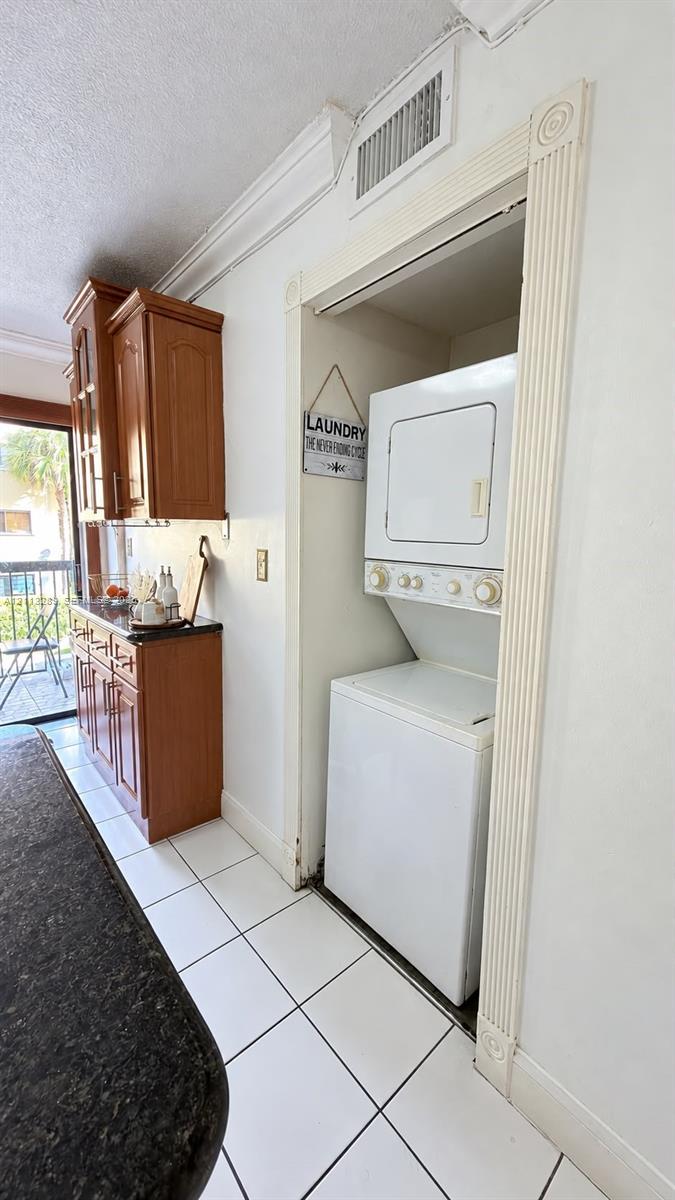 Stacked washer and dryer unit, wood cabinetry with stone-finish countertops, and white tile flooring