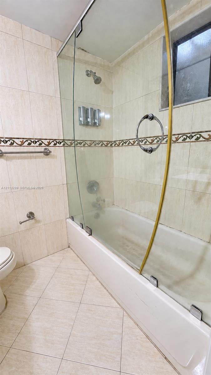 Bathtub with a glass shower enclosure, decorative tile accent border, wall-mounted metallic fixtures, and ceramic floor tiling