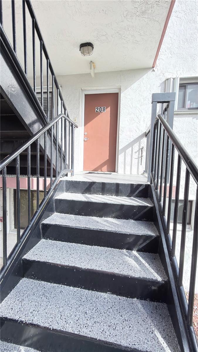 Exterior staircase featuring black metal railings and concrete-finish treads with a textured surface, leading to an elevated landing with a covered entryway and ceiling-mounted light fixture