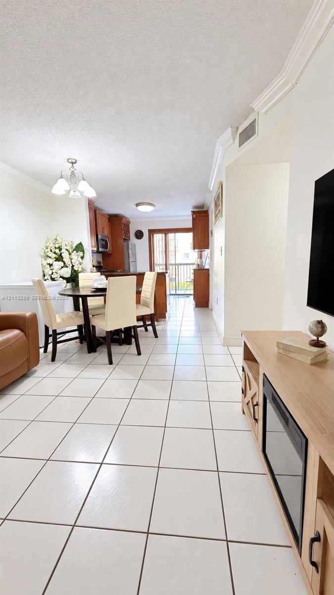 Open-concept interior featuring uniform white tile flooring, crown molding, wood-finish cabinetry, and glass sliding doors providing exterior access
