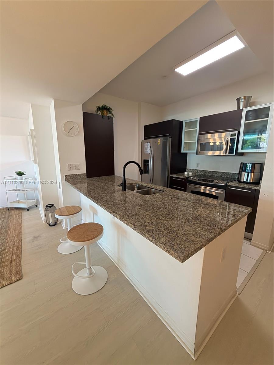 Kitchen featuring a peninsula with stone-finish countertops, stainless steel appliances, dark cabinetry, and a high-arc matte black faucet Kitchen featuring a peninsula with stone-finish countertops, stainless steel appliances, dark cabinetry, and a high-arc matte black faucet
