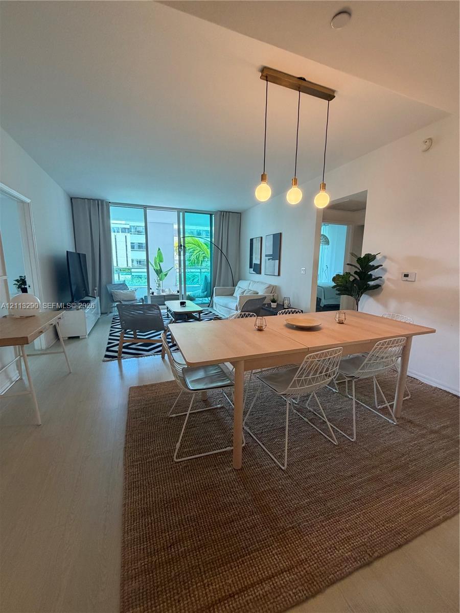 Open-concept living space featuring wood-finish flooring, floor-to-ceiling glass sliding doors, and a linear pendant light fixture with three exposed bulbs Open-concept living space featuring wood-finish flooring, floor-to-ceiling glass sliding doors, and a linear pendant light fixture with three exposed bulbs